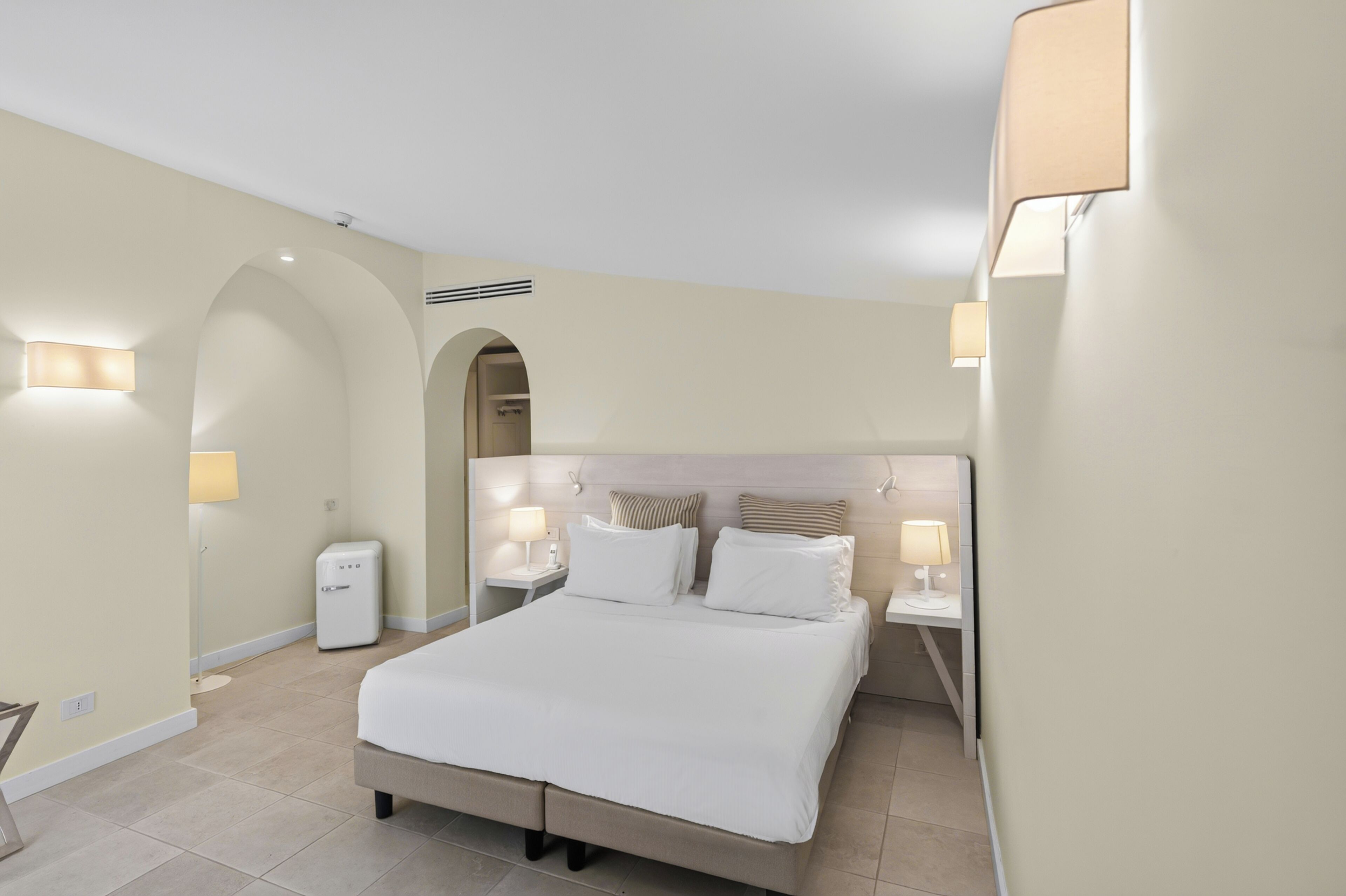 A hotel room with a large bed, two bedside tables with lamps, a small white appliance, and a wall-mounted heater.