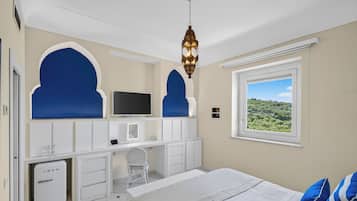 A bedroom with a bed, a desk, a chair, a television, a small refrigerator, and a window with a view.