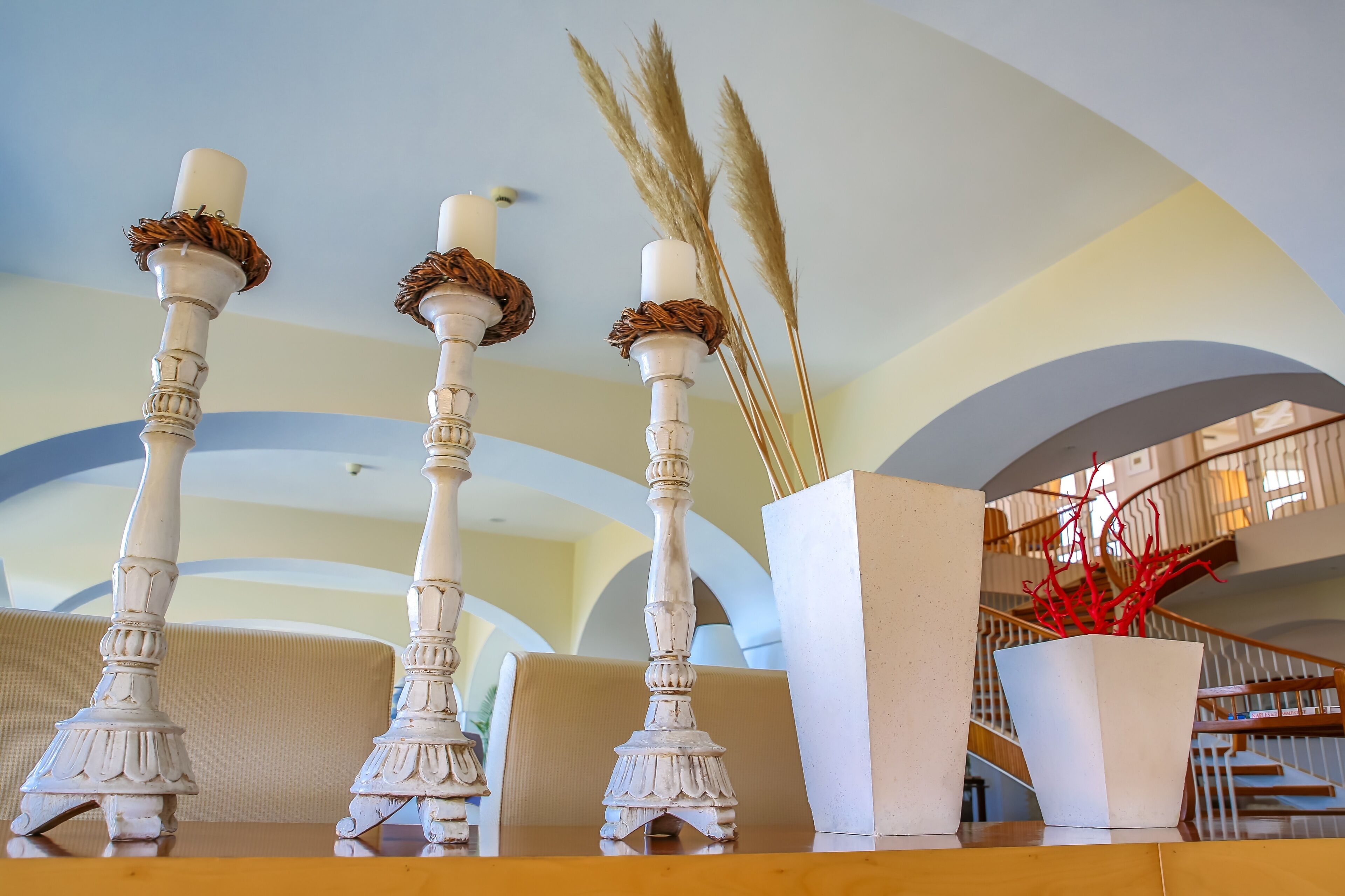 A room with three white candle holders with decorative elements, a white planter with red branches, and a staircase in the background.