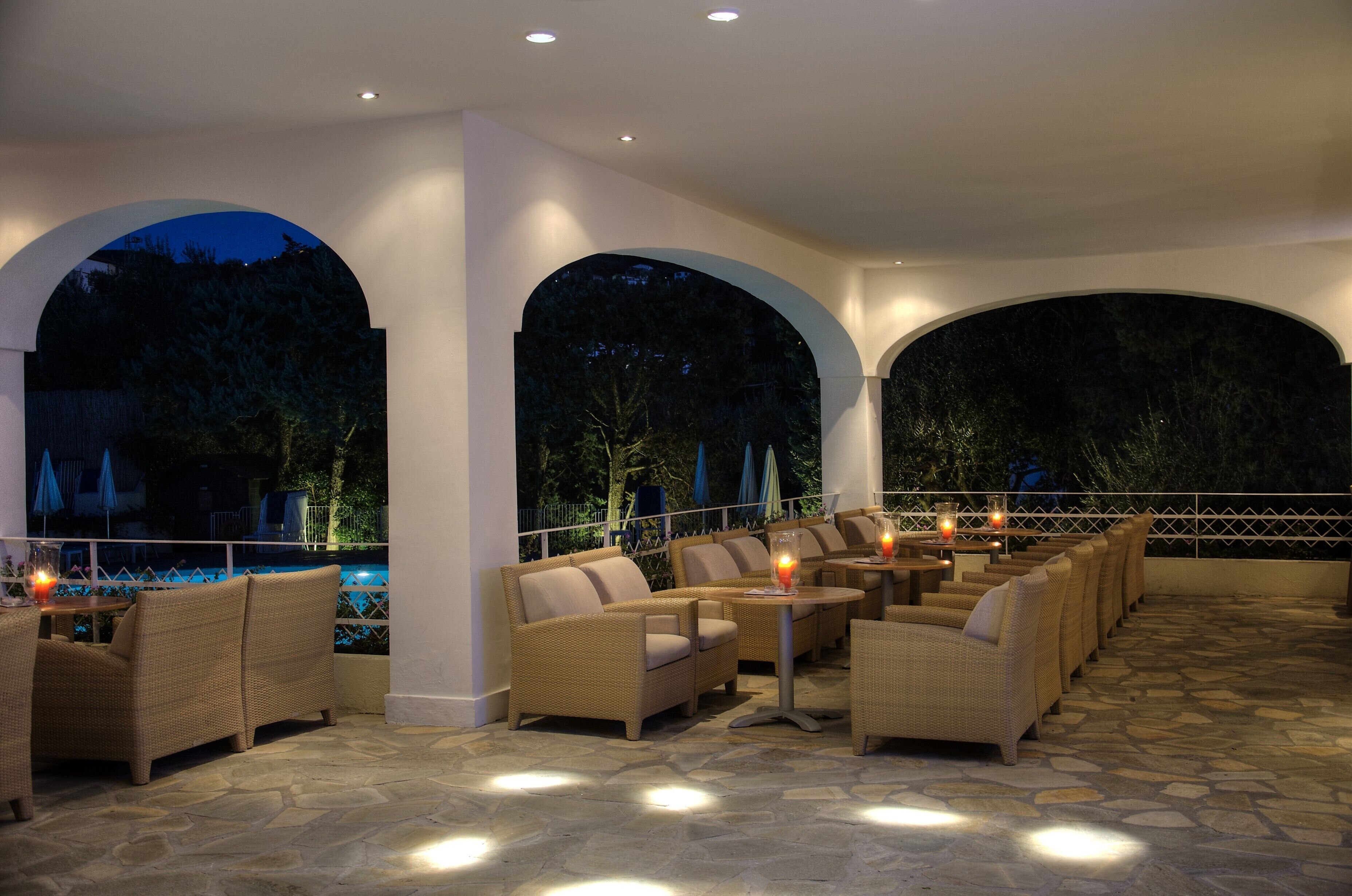 An outdoor patio area with wicker furniture, stone flooring, and a pool in the background.
