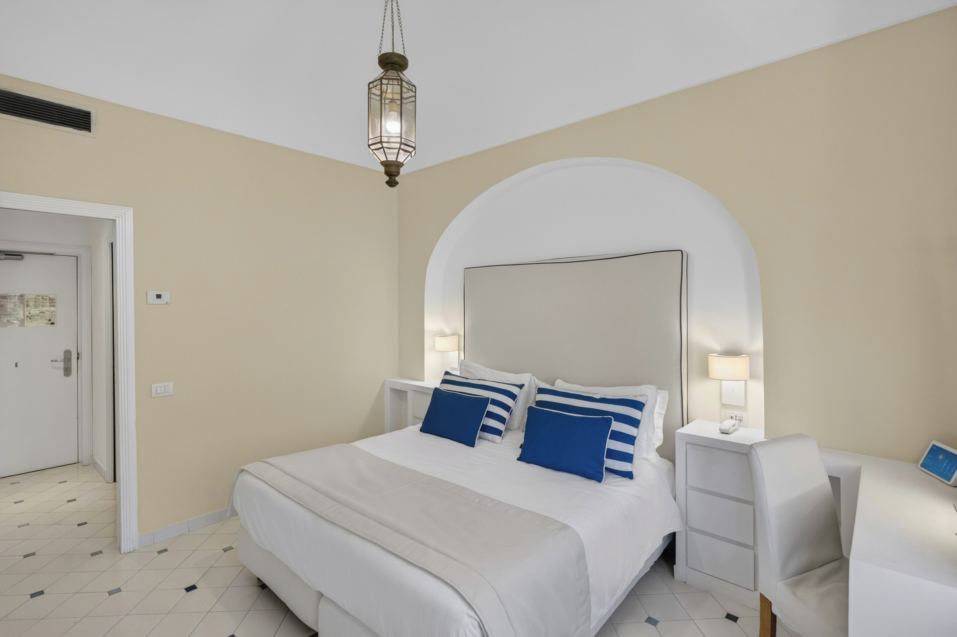 A hotel room with a large bed, bedside tables, a desk, and a hanging light fixture.