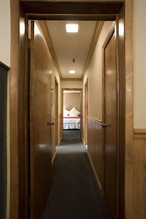 A hotel hallway with wooden doors, a carpeted floor, and a bed with a white bedspread and pillow in the background. - Alpenhof (Teton Village)
