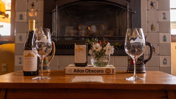 A wooden table with a wine bottle, two wine glasses, a candle, a book titled 'Atlas Obscura', and a decorative pitcher with flowers. - Alpenhof (Teton Village)