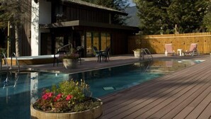 A wooden deck with a swimming pool, surrounded by tall trees and a building with a modern design. - Alpenhof (Teton Village)