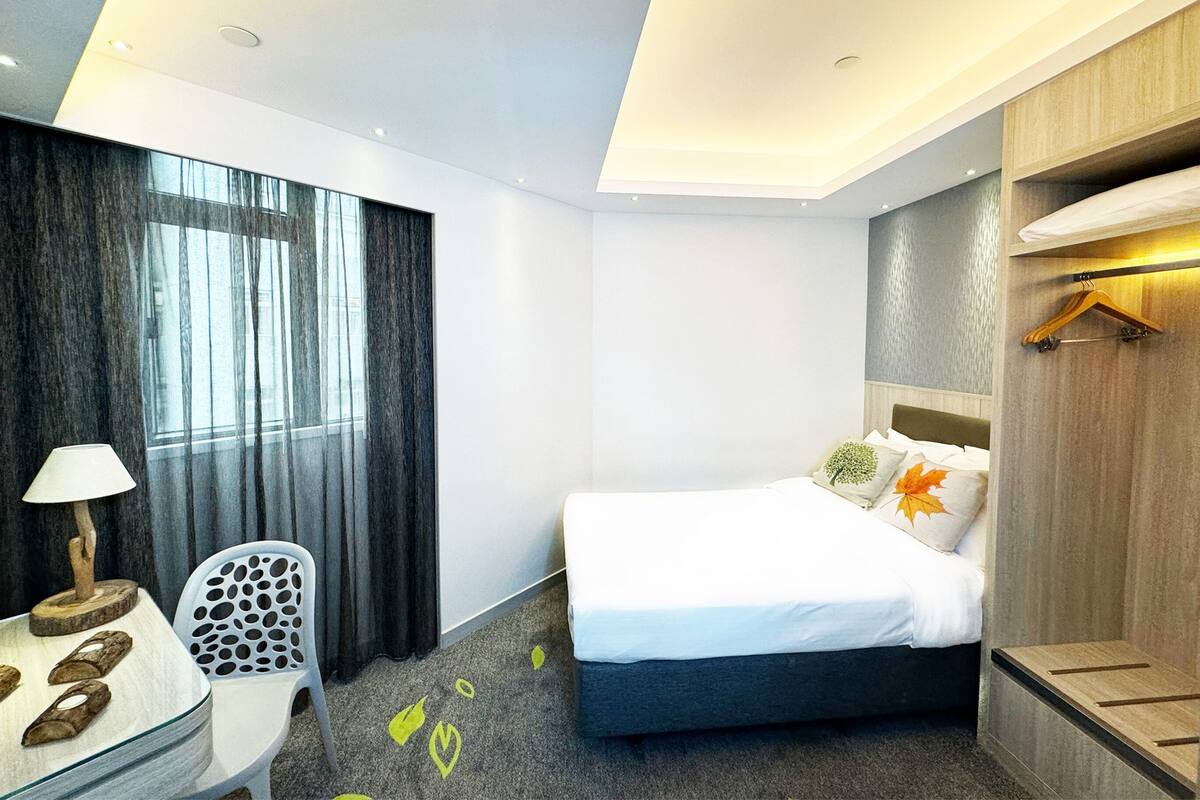 A modern hotel room with a bed, a desk with a lamp, a chair, and a wardrobe.