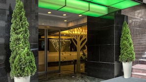 The entrance of the Stanford Hillview Hotel with a green-lit canopy, a glass door entrance, and two potted trees. - Stanford Hillview Hotel (Kowloon)