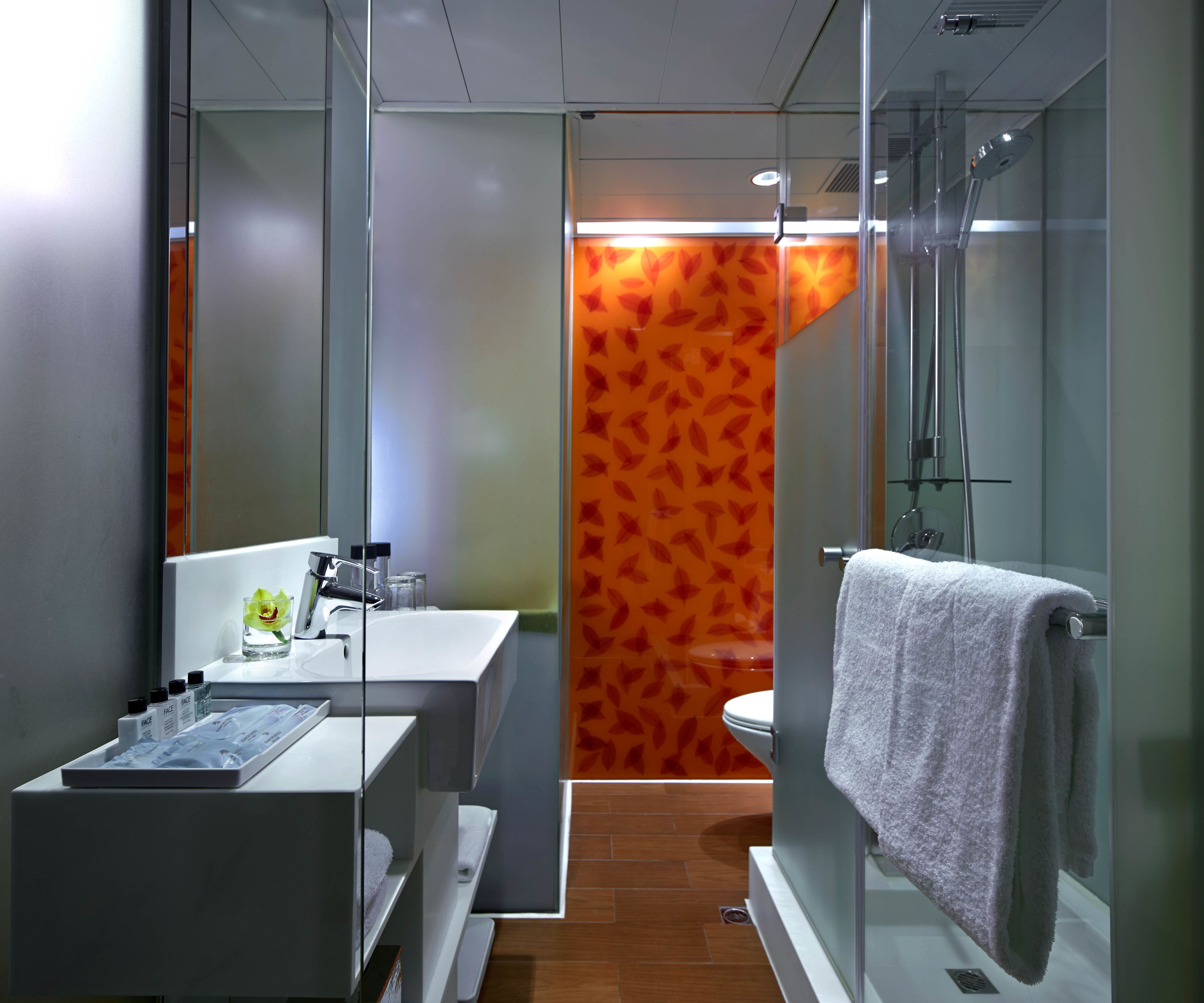 A modern bathroom with a glass shower enclosure, a white sink, and a toilet with an orange leaf-patterned wall behind it.