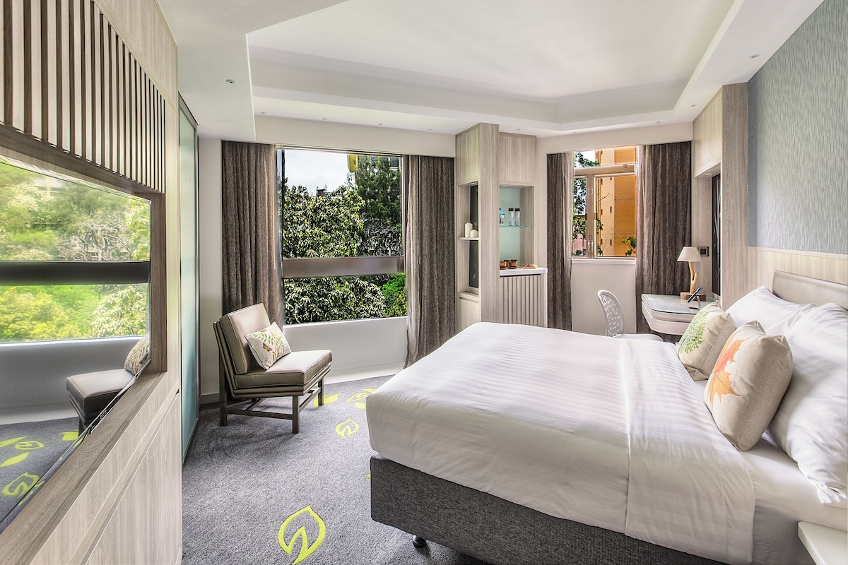 A modern hotel room with a large bed, a seating area with a chair and ottoman, a vanity with a mirror, and a window with a view of greenery.