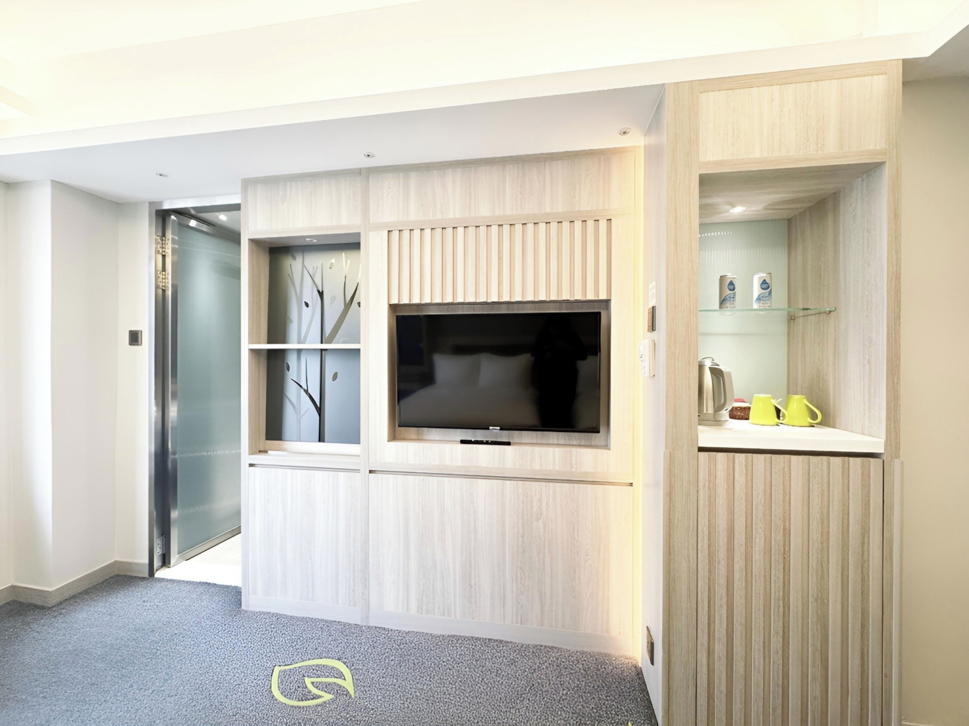 A modern hotel room with a built-in TV, a glass door leading to a balcony, and a small kitchenette area.