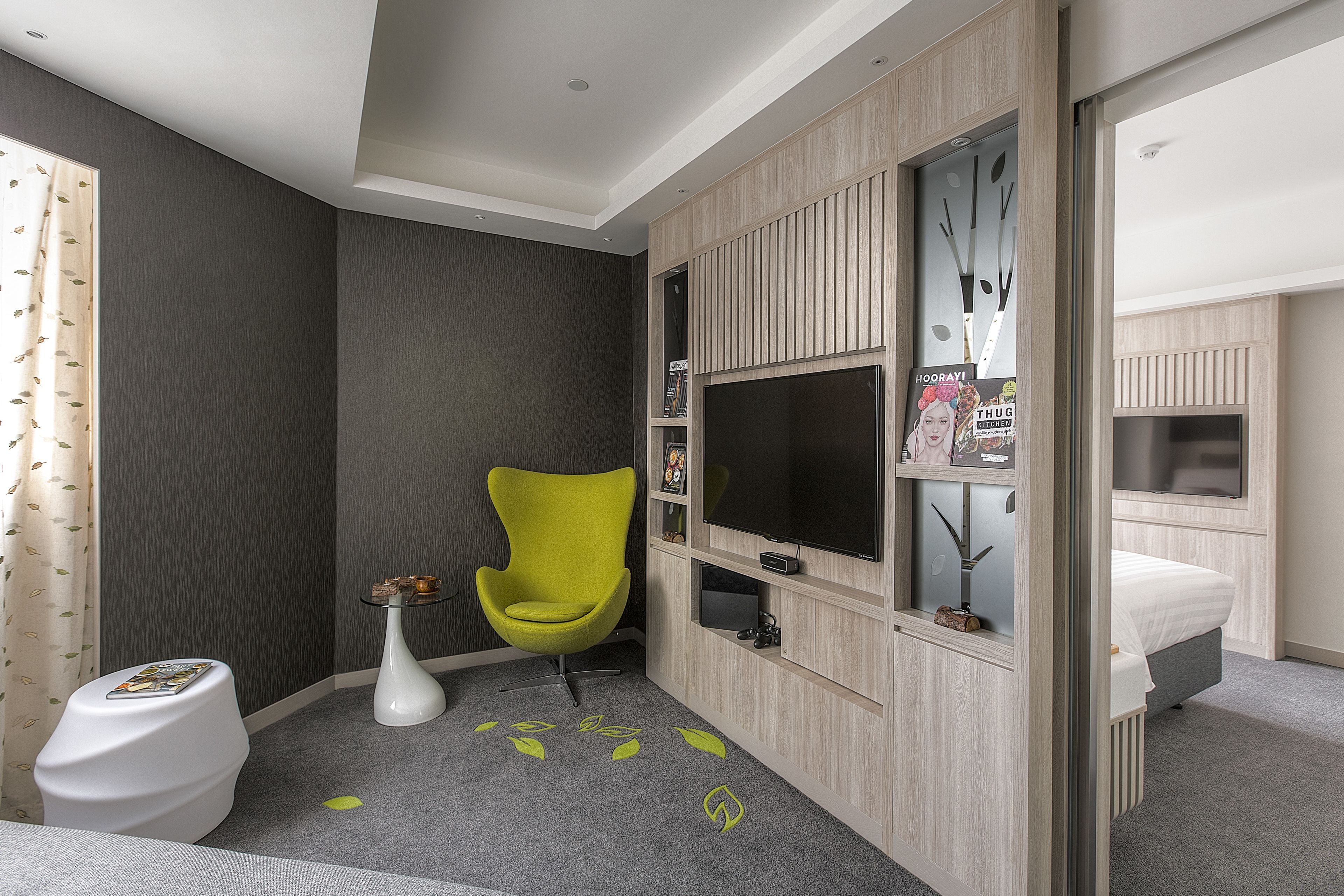 A modern hotel room with a grey carpet, a bright yellow chair, a white toilet, and a bed with a grey headboard.