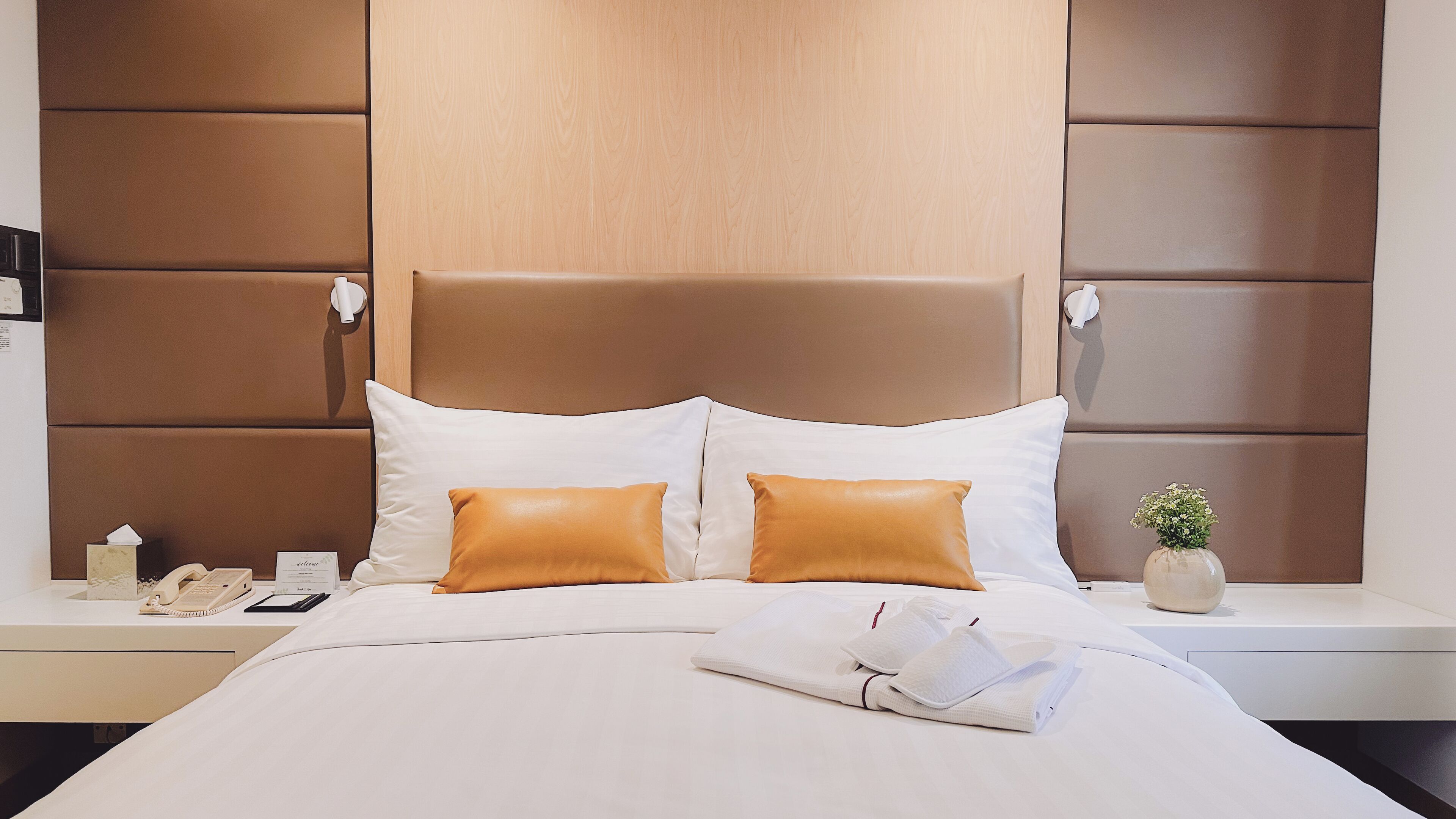A hotel room with a large bed, two pillows, a bedside table with a lamp, a vase with flowers, and a small plant.