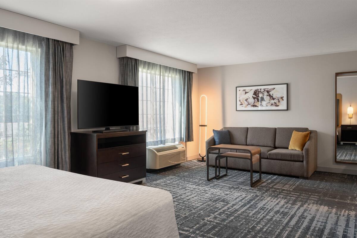 Una camera d'hotel con un letto, una TV a schermo piatto, un divano, un tavolino da caffè, una lampada e un quadro appeso al muro.