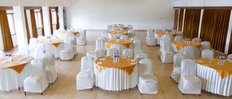 A large conference room with multiple round tables covered with white tablecloths and orange centerpieces, surrounded by white chairs.