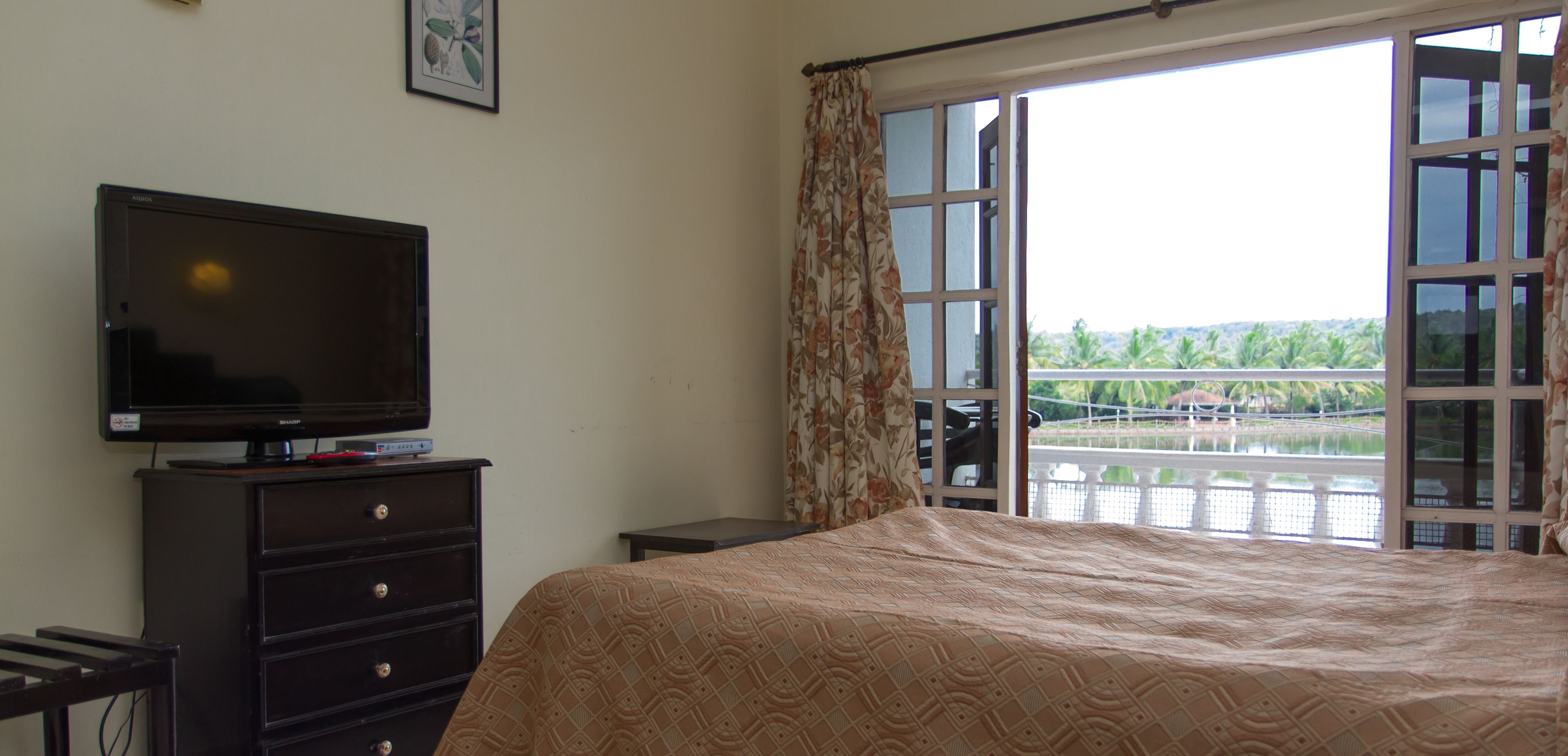 Quarto de hotel com uma cama, televisão em uma cômoda e uma grande janela com vista para um corpo d'água e vegetação.