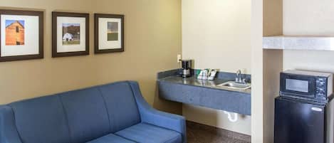 Ein Hotelzimmer mit blauem Sofa, einer Küchenzeile inklusive Mikrowelle, Kaffeemaschine und Spüle. An der Wand hängen drei gerahmte Bilder.