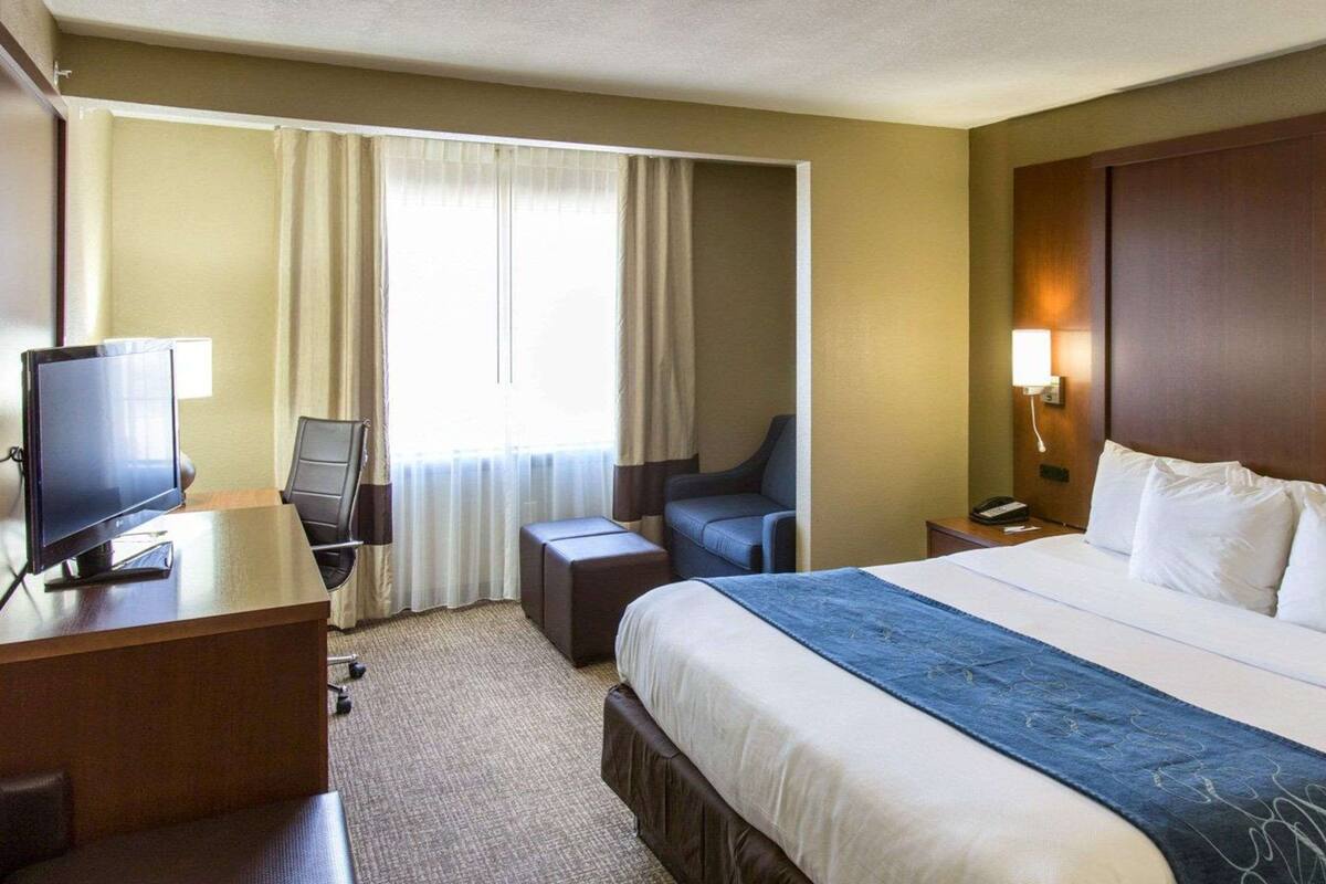 A hotel room with a bed, a desk with a TV, a chair, a small table, and a nightstand with a lamp.