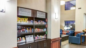 A hotel lobby with a self-service kiosk labeled 'Your Suite Success Marketplace' stocked with snacks and beverages. There are seating areas with blue chairs and a wooden coffee table. - Comfort Suites Idabel (Idabel)