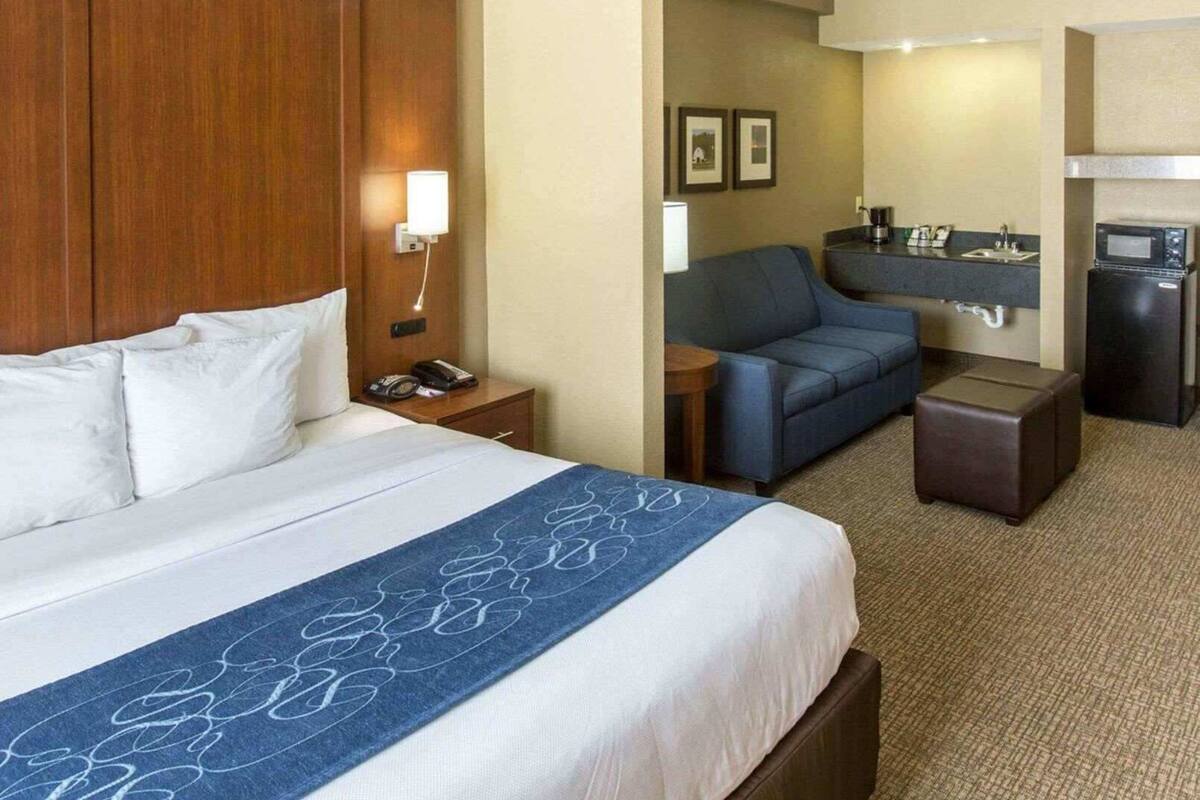 A hotel room with a bed, a desk, a chair, a small table, a microwave, and a refrigerator.