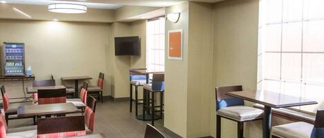 A hotel room with a dining area featuring tables and chairs, a flat-screen TV mounted on the wall, and a vending machine.