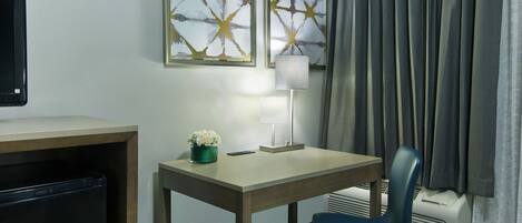 A hotel room with a desk, chair, lamp, and artwork on the wall.