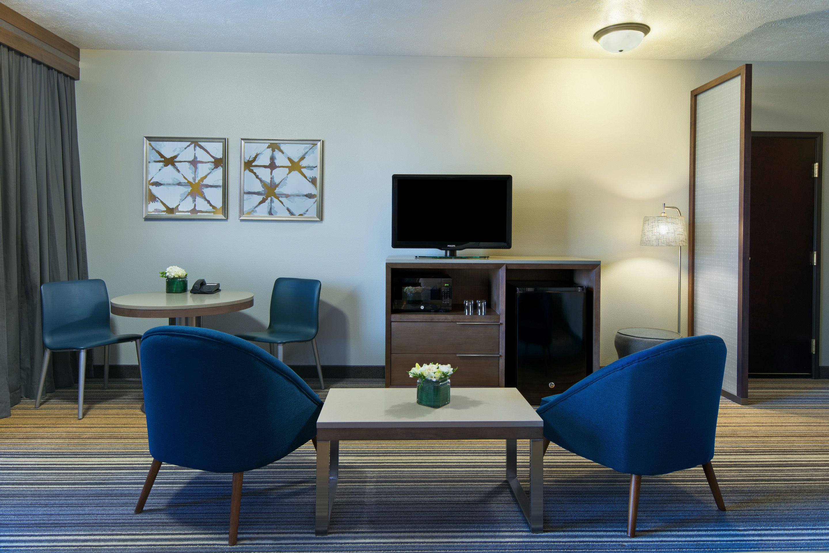 Habitación de hotel con televisión, dos sillas azules, una mesita con un florero y una obra de arte en la pared.