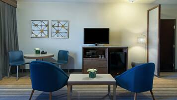 Habitación de hotel con televisión, dos sillas azules, una mesita con un florero y una obra de arte en la pared.