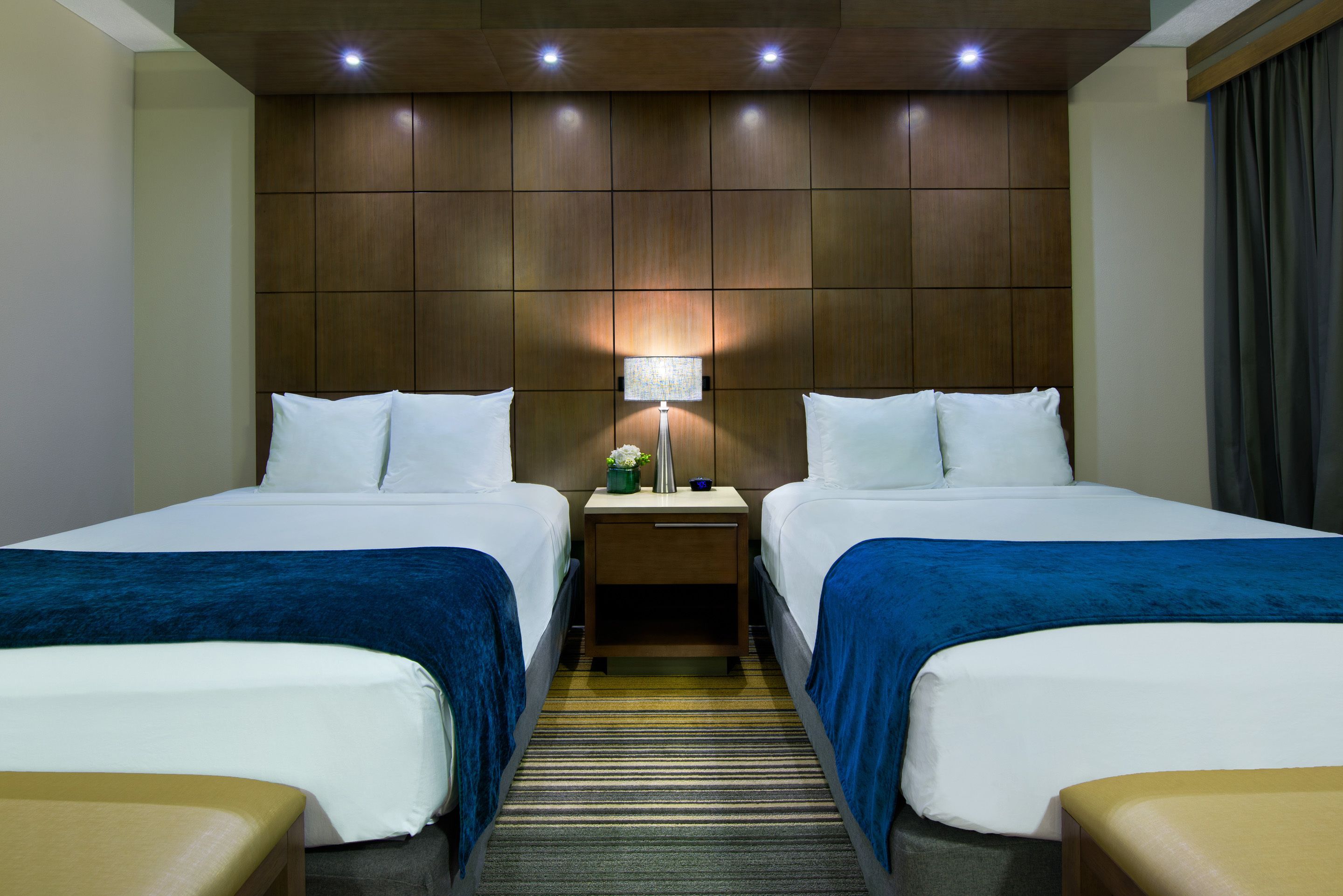 A hotel room with two beds, a nightstand with a lamp, and a striped carpet.