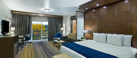 A hotel room with a large bed, a desk with a computer, a chair, a small table, and a view of the outdoors.