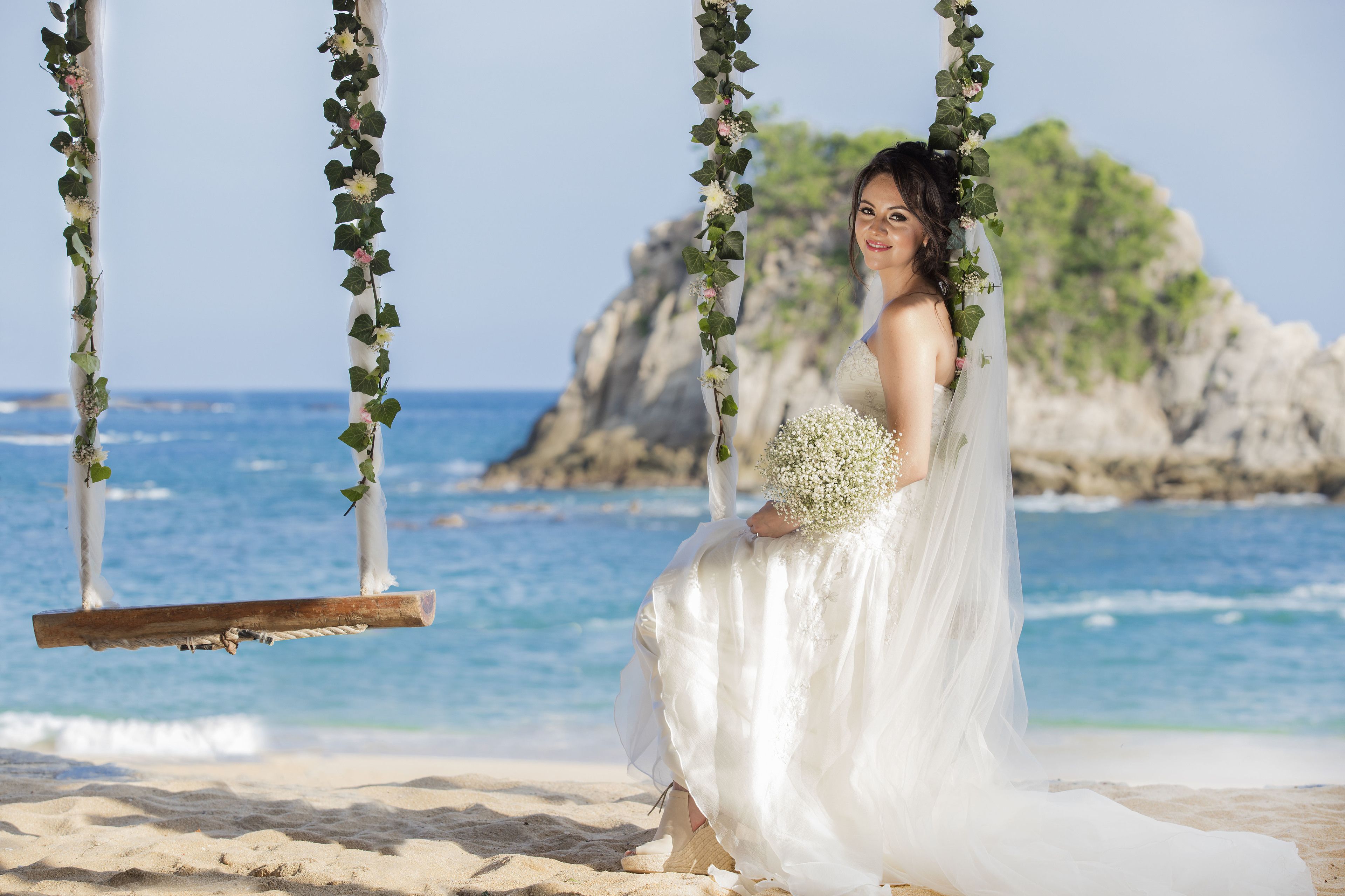 A wedding scene on a beach with a swing decorated with greenery and flowers, a bride in a white dress, and a rocky cliff in the background.