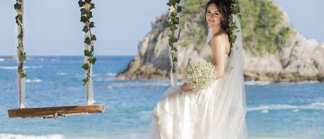 A wedding scene on a beach with a swing decorated with greenery and flowers, a bride in a white dress, and a rocky cliff in the background.