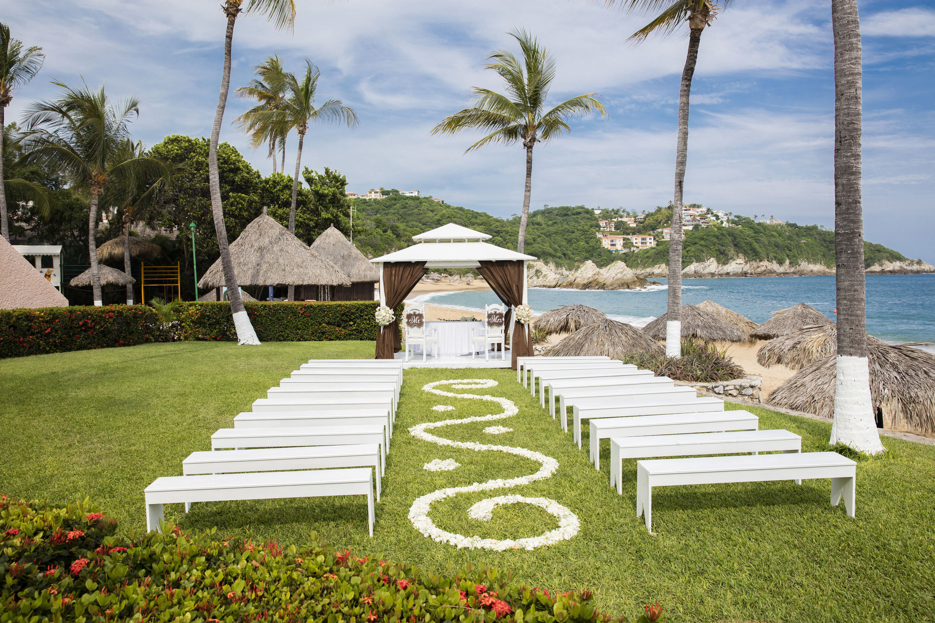 A beachside wedding setup with a white aisle, benches, and a gazebo.