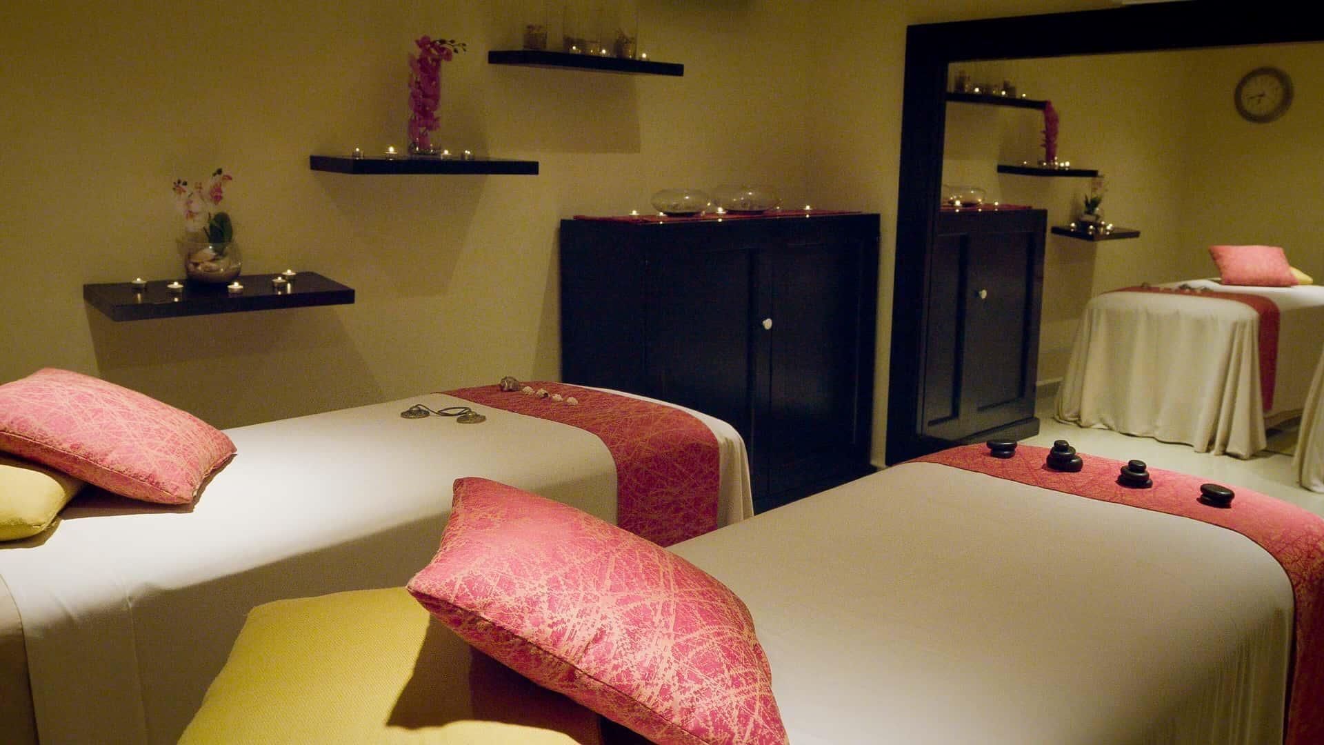 Un cuarto de spa con dos camas, cada una con un almohadón rojo y amarillo, y un conjunto de piedras negras sobre la cama. Hay repisas con velas y un espejo en la pared.