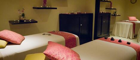 Un cuarto de spa con dos camas, cada una con un almohadón rojo y amarillo, y un conjunto de piedras negras sobre la cama. Hay repisas con velas y un espejo en la pared.