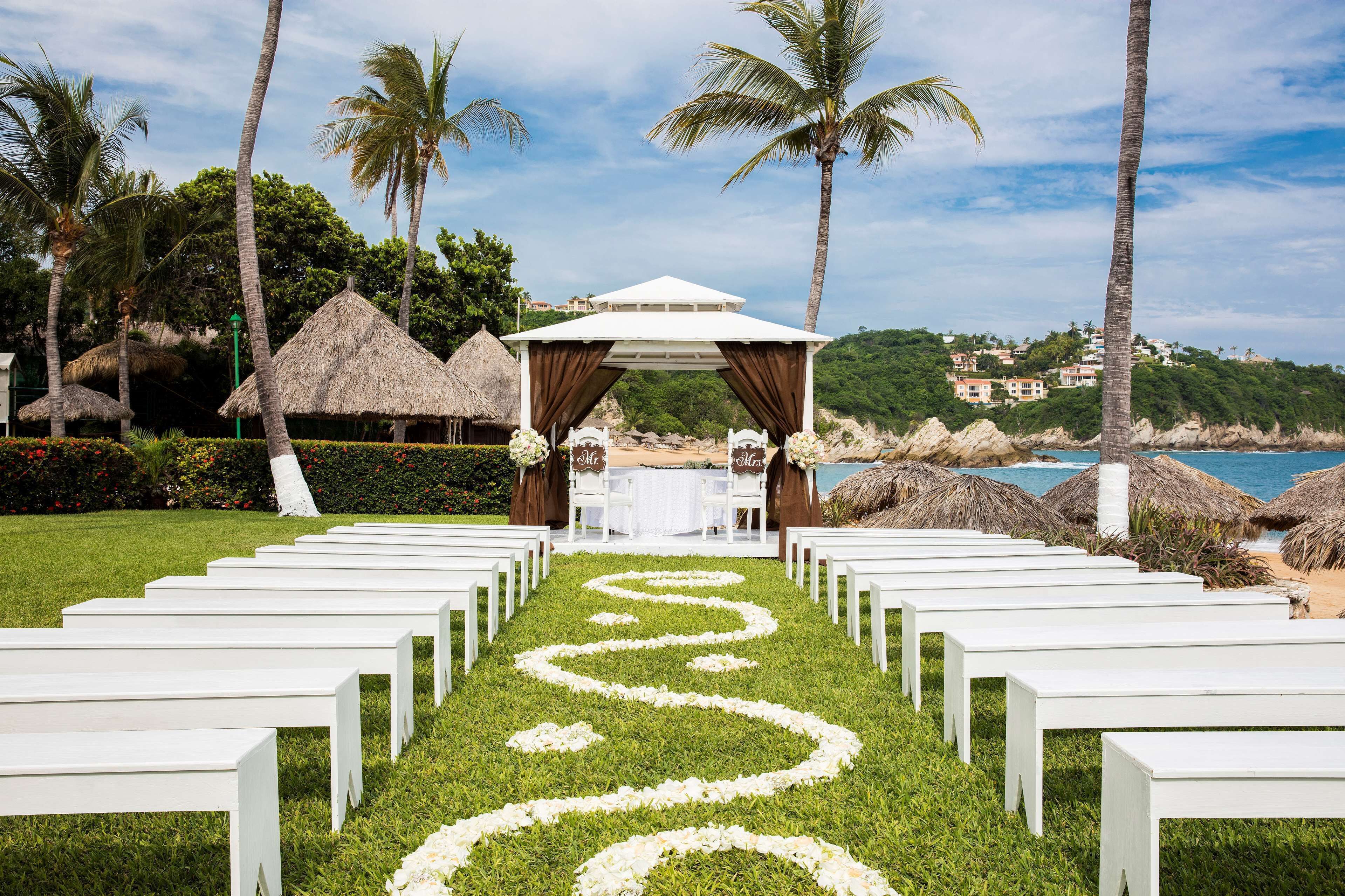 Decoración para una boda en la playa, con un pasillo blanco, un pabellón y chozas de techo de paja.
