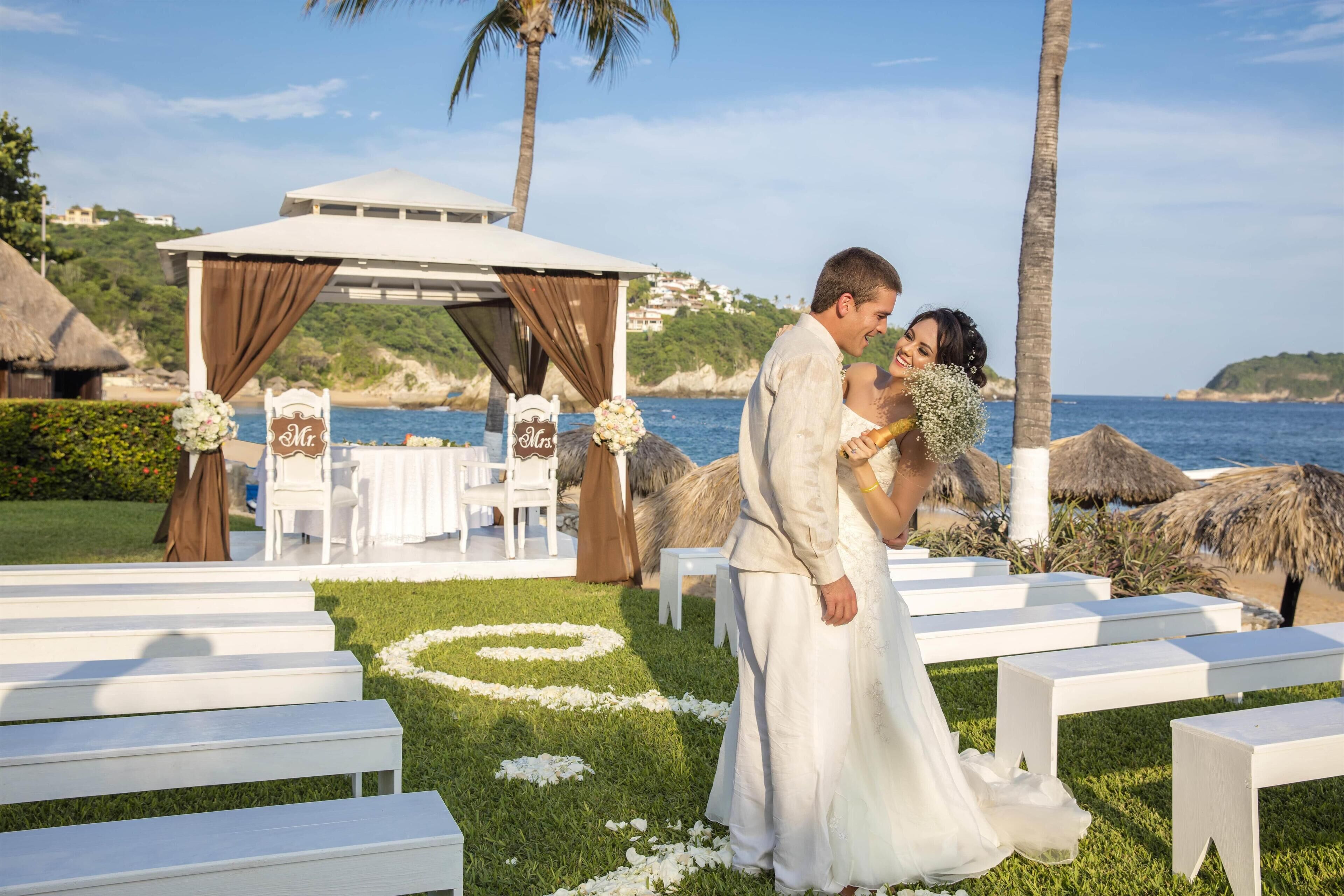 A beachside wedding ceremony with a white gazebo, white benches, and a sea view.