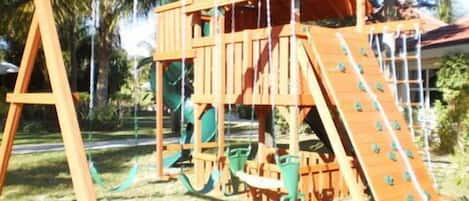 Estructura de madera con tobogán y red de escalada, ubicada en un área verde con palmeras.