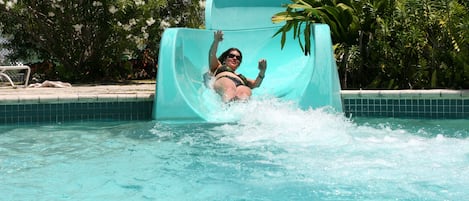 A person sliding down a turquoise water slide into a pool surrounded by lush greenery.