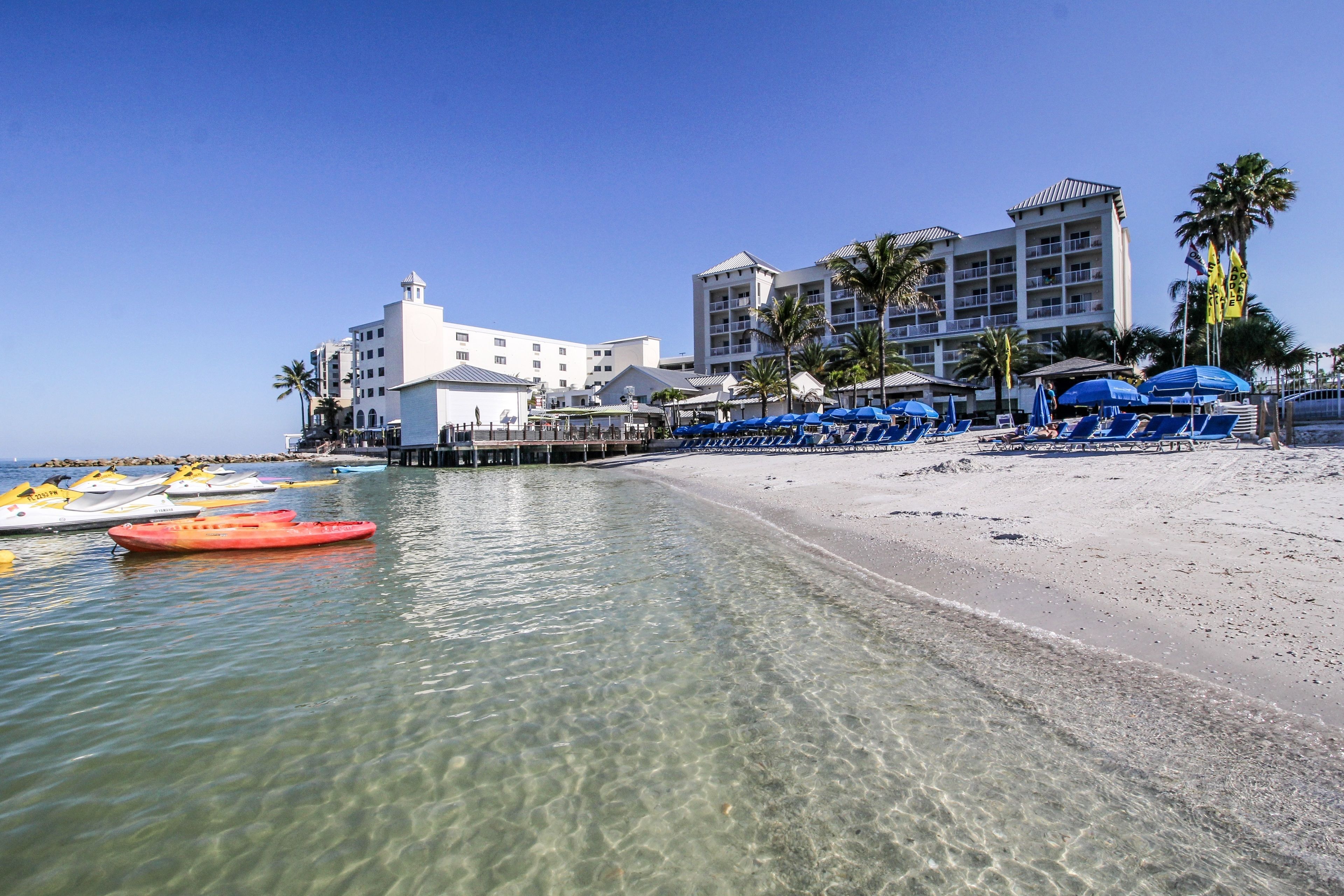 Een strand met helder water, een rij blauwe strandstoelen en een hotel met meerdere verdiepingen en balkons.