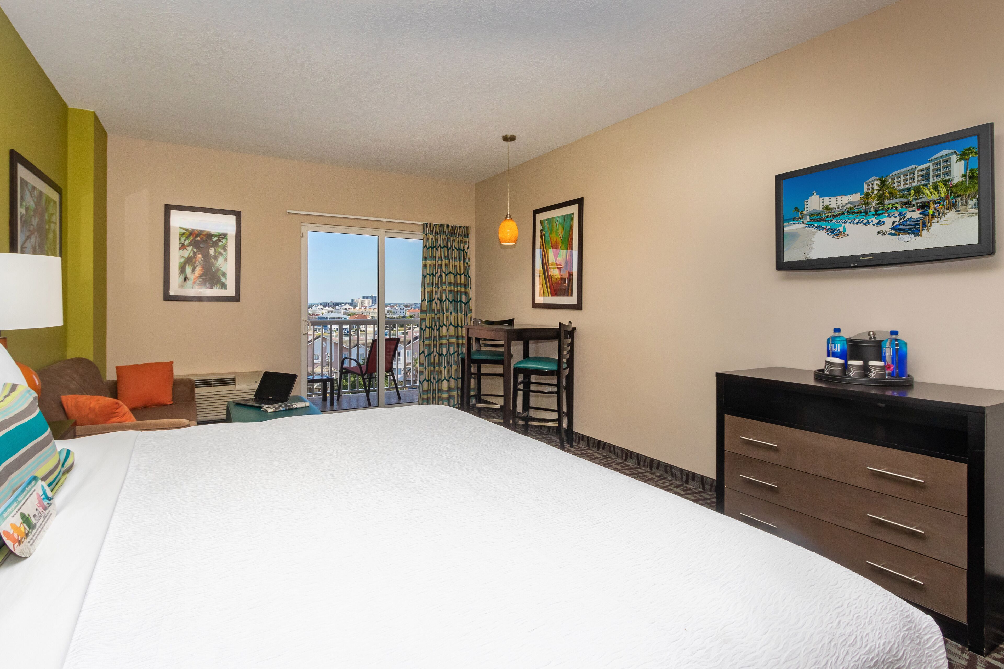 A hotel room with a large bed, a desk with a laptop, a TV, and a balcony with a view of the city.