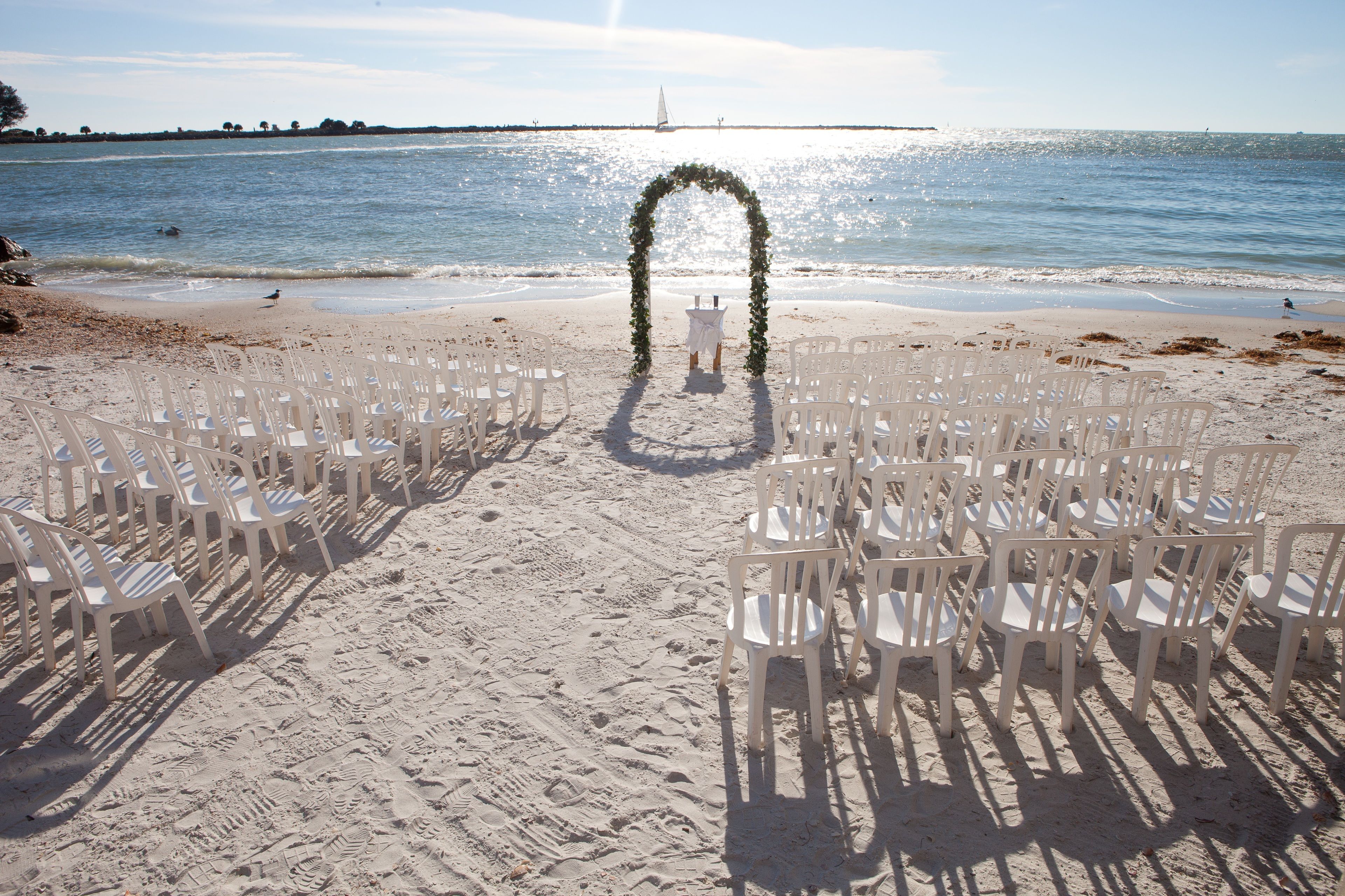 Decoración para una boda en la playa con sillas de plástico blancas dispuestas en filas, un arco blanco y un velero al fondo.