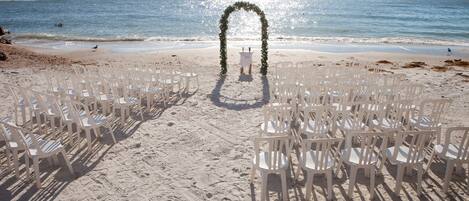 Decoración para una boda en la playa con sillas de plástico blancas dispuestas en filas, un arco blanco y un velero al fondo.