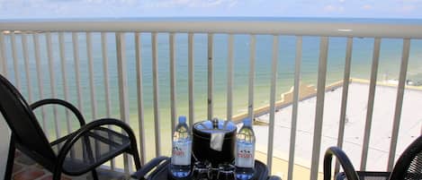 A balcony with a view of the ocean, two chairs, a table with bottles and glasses, and a pot.