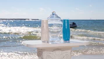 Una mesa con un tarro de vidrio, un vaso y un vaso azul en una playa, con olas al fondo.