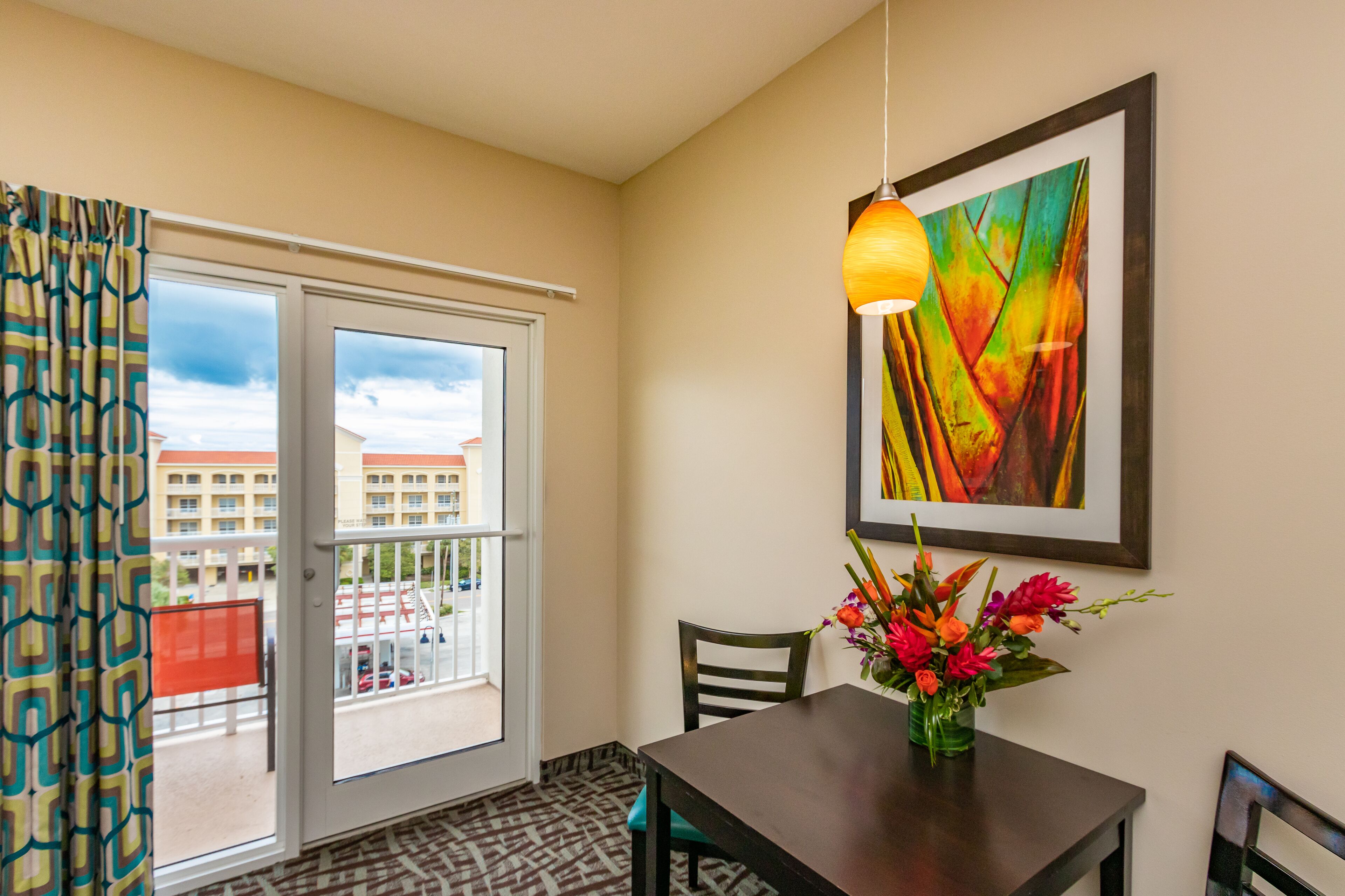 Une chambre d’hôtel avec une table à manger, des chaises, un vase de fleurs, un tableau abstrait encadré et une vue sur les bâtiments à travers une porte vitrée.