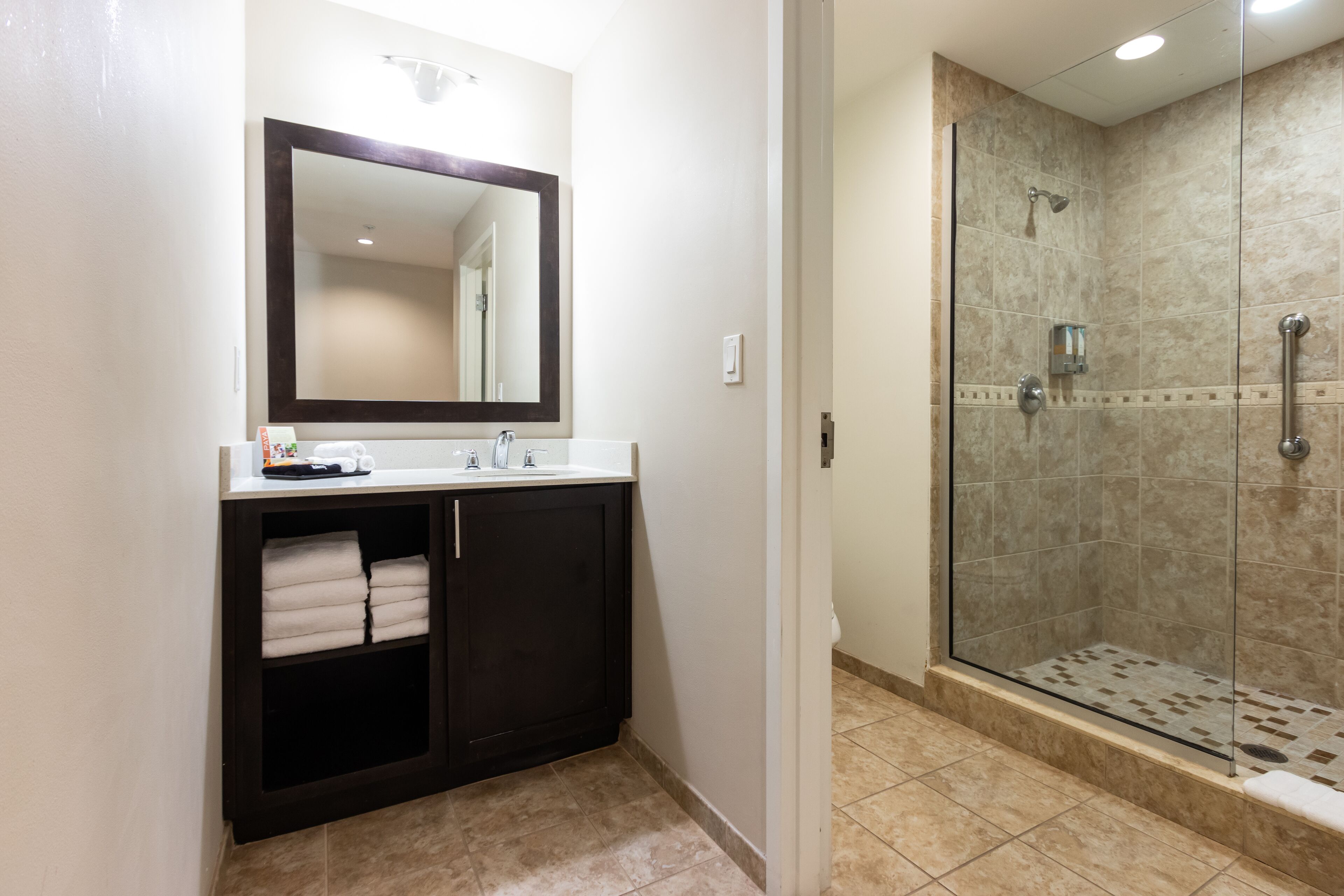 Un baño con tocador de madera oscura, un espejo grande, cabina de ducha con mampara de vidrio y suelo de baldosas beige.