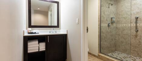 Un baño con tocador de madera oscura, un espejo grande, cabina de ducha con mampara de vidrio y suelo de baldosas beige.
