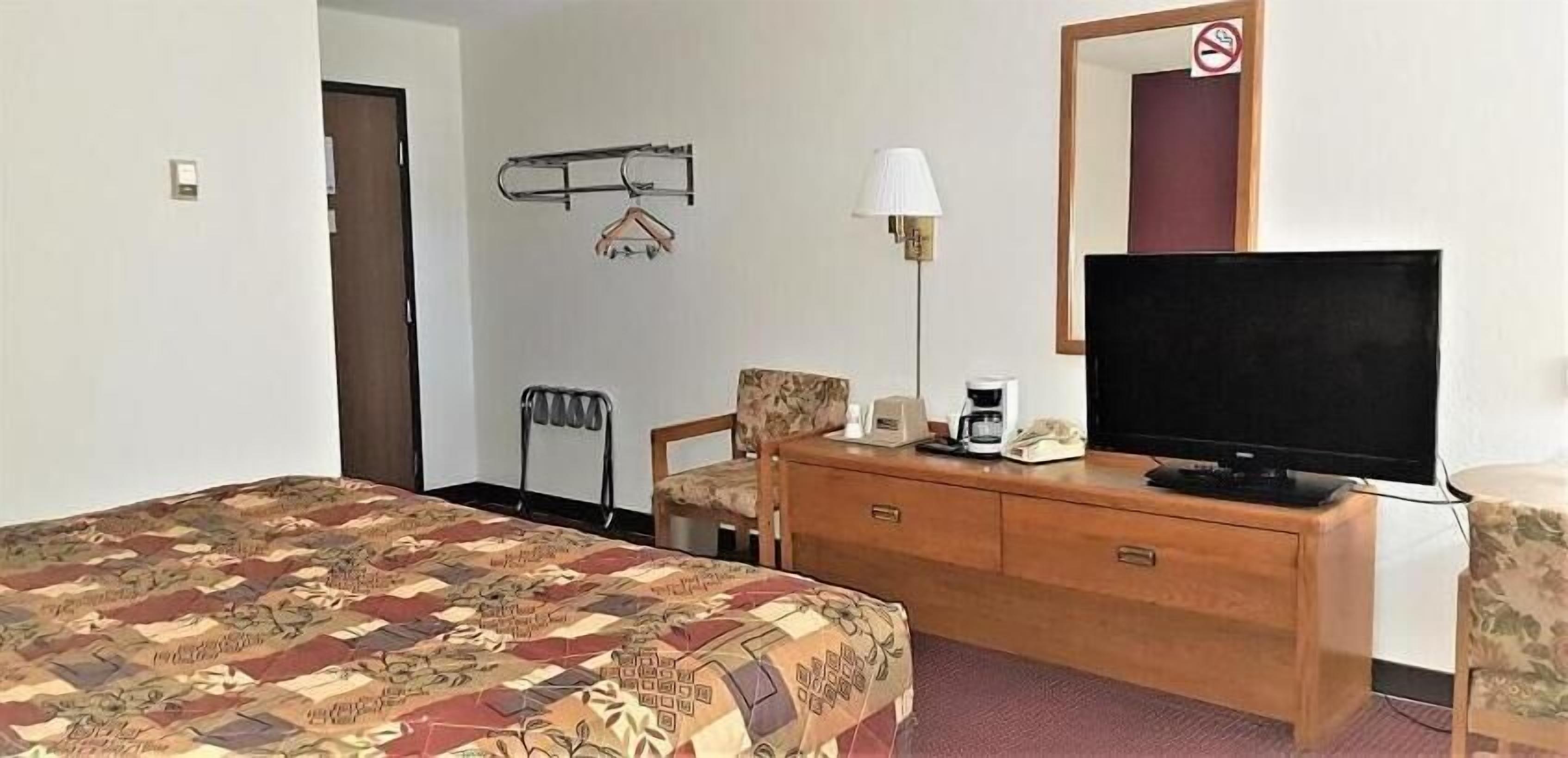 A hotel room with a bed, a wooden dresser, a television, a chair, a desk, and a lamp.