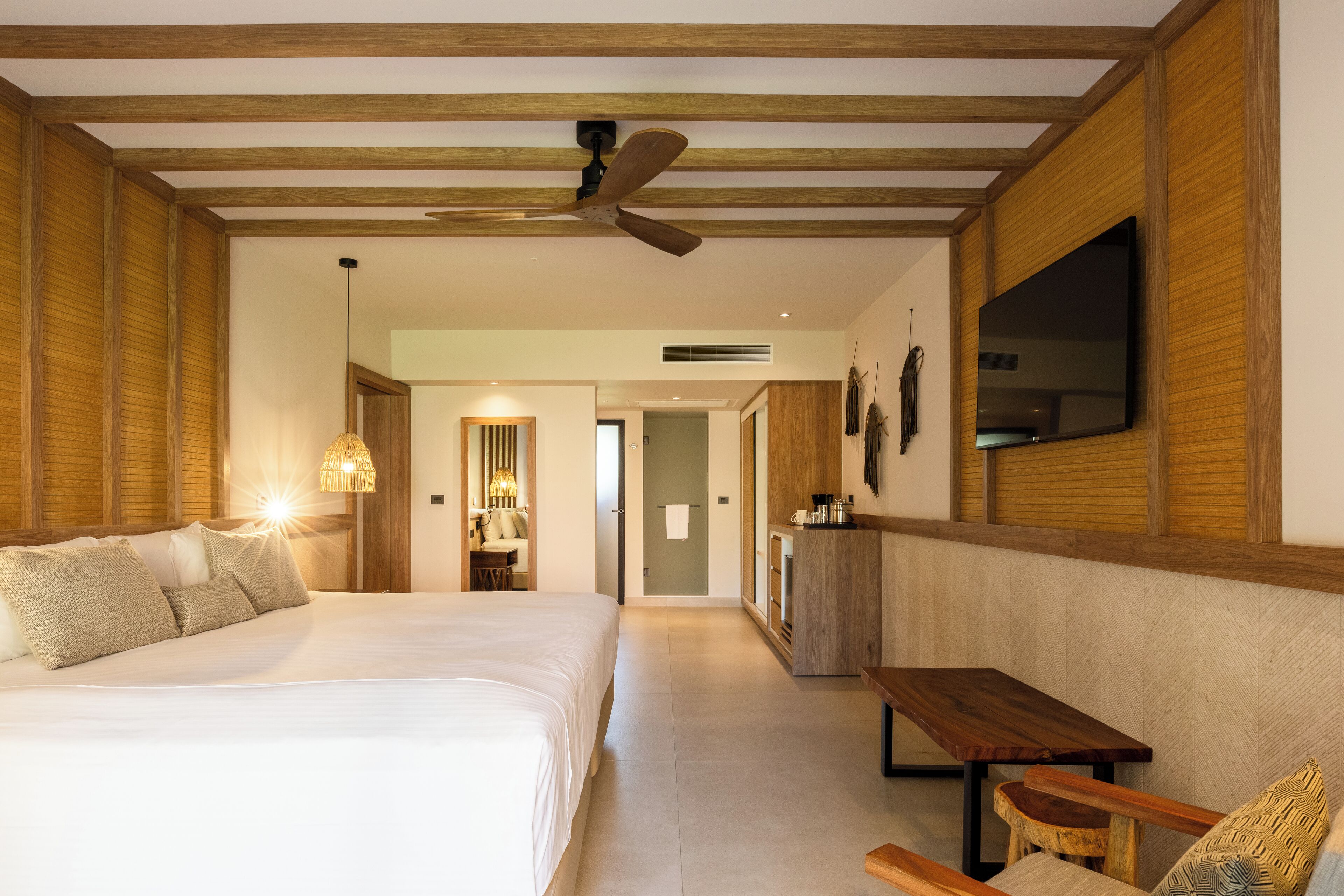A hotel room with a large bed, a wooden ceiling, a ceiling fan, a TV mounted on the wall, a small table, and a bench.