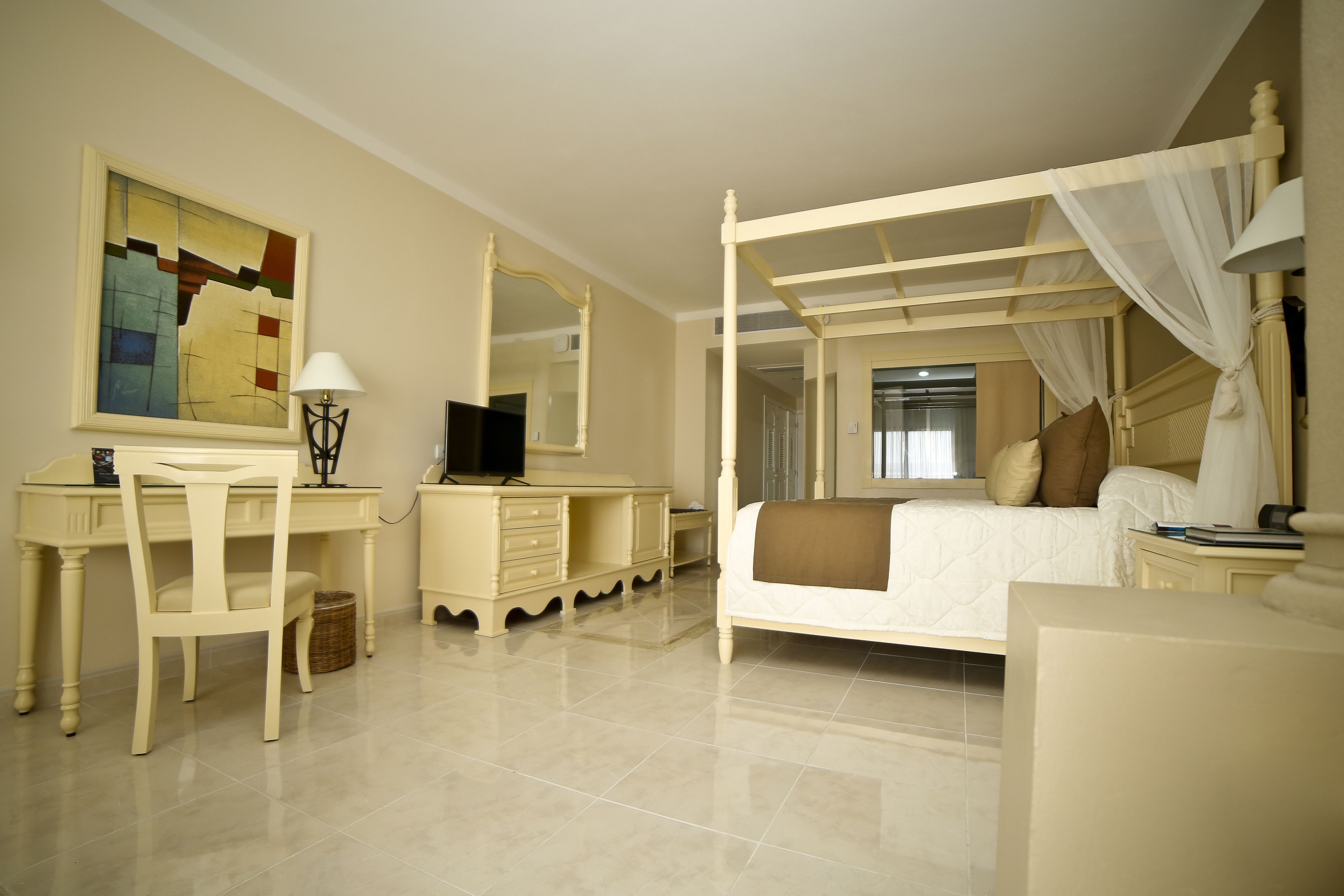A bedroom with a four-poster bed, a desk with a chair, a television, and a large painting on the wall.