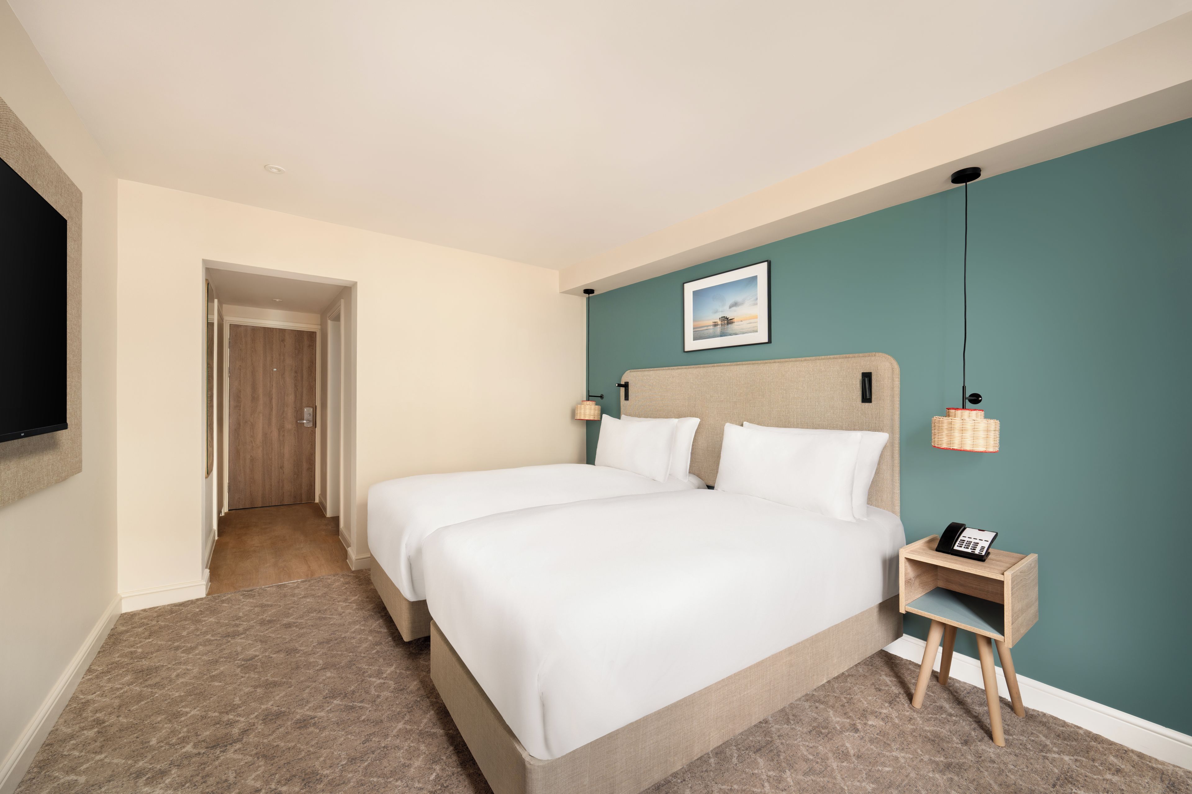 A hotel room with two beds, a small bedside table with a phone, a wall-mounted picture, and a door leading to another room.
